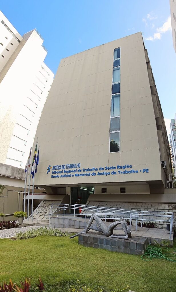 Fachada do prédio do Memorial e da Escola Judicial do TRT6, em Pernambuco. Edifício alto, de cor clara, com janelas verticais no lado direito. Na frente, há gramado, mastros com bandeiras, uma escultura de figura humana reclinada e rampa de acesso com corrimão. Céu azul ao fundo.