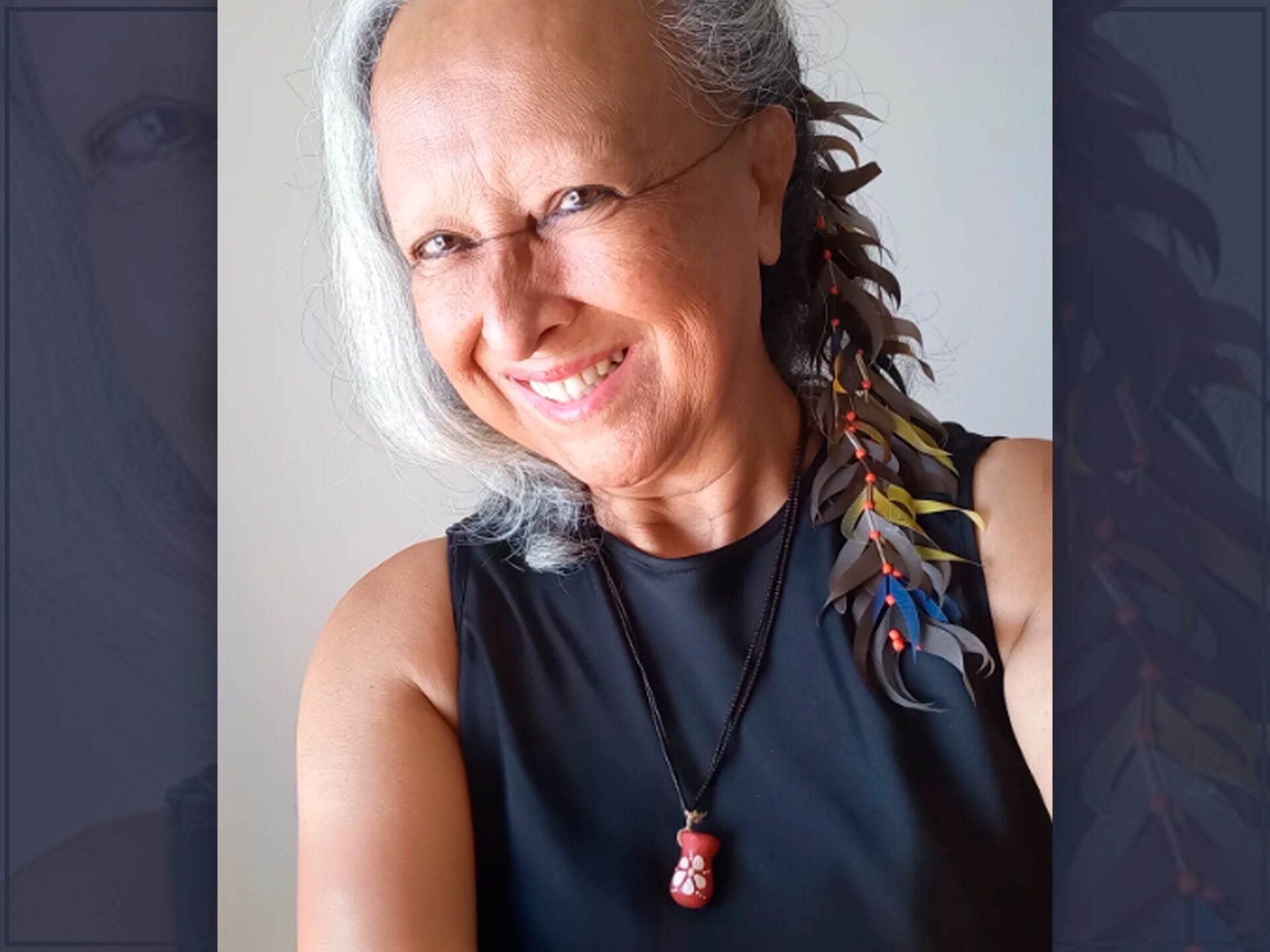 Fotografia de Shirley Vilhalva, mulher indígena surda de cabelos grisalhos adornados com trança de folhas coloridas e sementes vermelhas. Veste regata preta e colar com pingente de cerâmica vermelho. Sorri levemente de frente para a câmera.