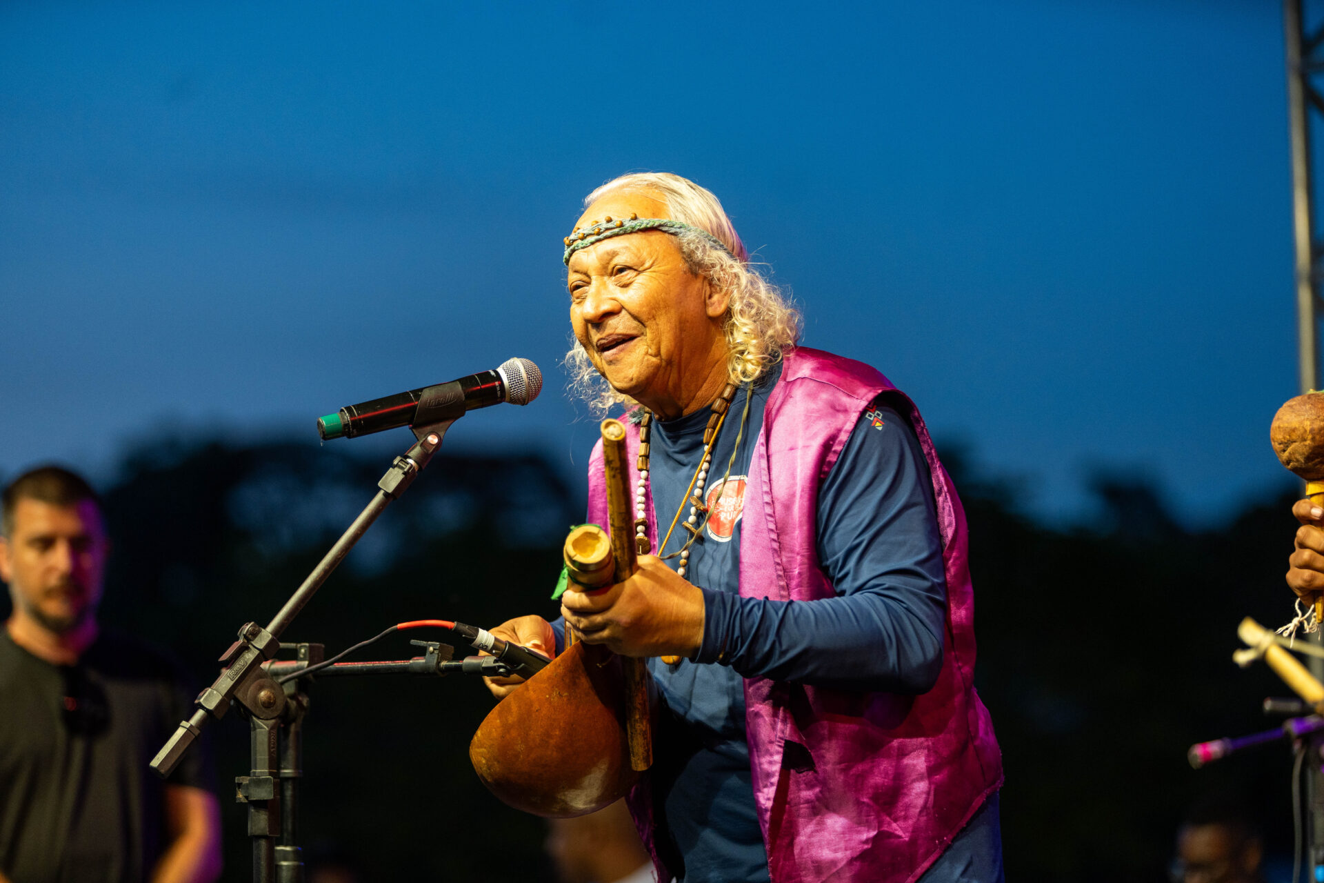 Fotografia de Dauá Puri, homem indígena de cabelos brancos, sorrindo ao microfone. Segura instrumento de cabaça, usa colete roxo e adereços tradicionais. Fundo: céu azul ao entardecer em um palco.