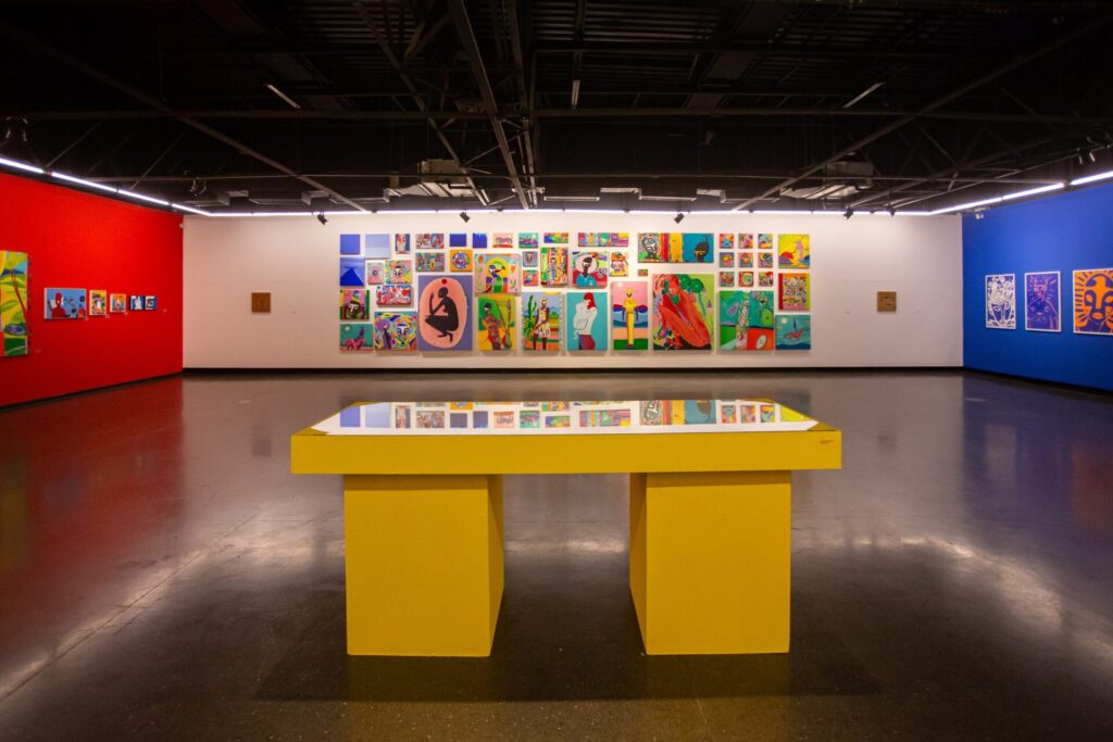 Sala ampla de museu ou galeria, com um espaço aberto e organizado. No centro, há uma mesa retangular grande, pintada de amarelo, com tampo de vidro. Ao fundo, uma longa parede branca exibe dezenas de quadros coloridos organizados em uma grade. As obras apresentam diferentes estilos, muitas com figuras humanas, padrões abstratos e cores vibrantes. As paredes laterais são pintadas em cores fortes: vermelho à esquerda e azul à direita. O teto é escuro, com estruturas aparentes e iluminação linear. O chão também é escuro e liso, contribuindo para o contraste com as cores vivas das obras.