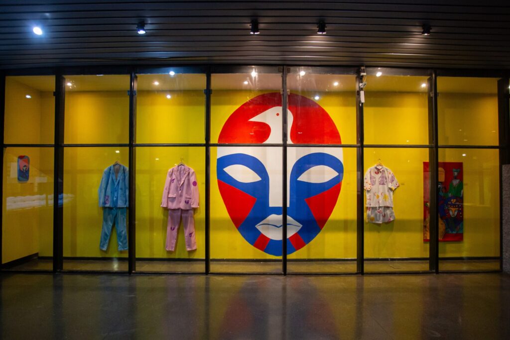 Ambiente interno de exposição, visto de frente. Há uma grande parede amarela ao fundo, protegida por uma estrutura de vidro com divisões pretas, como se fosse uma vitrine ampla. No centro, destaca-se um grande rosto estilizado, com formas geométricas em vermelho, azul e branco, lembrando uma máscara artística. Nas laterais desse rosto, estão expostos conjuntos de roupas penduradas: à esquerda, um terno azul claro e outro rosa; à direita, uma camisa e um conjunto com estampas coloridas. As peças parecem fazer parte de uma exposição de moda ou arte contemporânea. A iluminação é artificial, com focos de luz no teto. O chão é escuro e polido, refletindo levemente o ambiente.