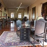 Uma fotografia de plano médio mostra o interior de uma sala histórica em um museu. O ambiente possui um teto alto e plano, paredes em tons de creme com detalhes ornamentais na base e um piso de madeira polida com tábuas largas e escuras. No centro do primeiro plano, há uma escrivaninha de madeira escura com gavetas entalhadas, acompanhada por cadeiras de espaldar alto com detalhes em madeira trabalhada. Sob o conjunto de móveis, estende-se um tapete oriental grande com padrões intrincados em tons de azul, bege e vermelho. Ao longo das paredes, diversos itens históricos estão expostos: À esquerda: Um armário escuro e alto, seguido por uma armadura medieval completa montada em um suporte. À direita: Uma cristaleira de madeira alta com portas de vidro e quadros emoldurados pendurados na parede Ao fundo: Duas janelas em arco permitem a entrada de luz natural, e um lustre trilho moderno no teto ilumina o corredor central da sala. A atmosfera é solene e preservada, evocando um estilo colonial ou clássico europeu.