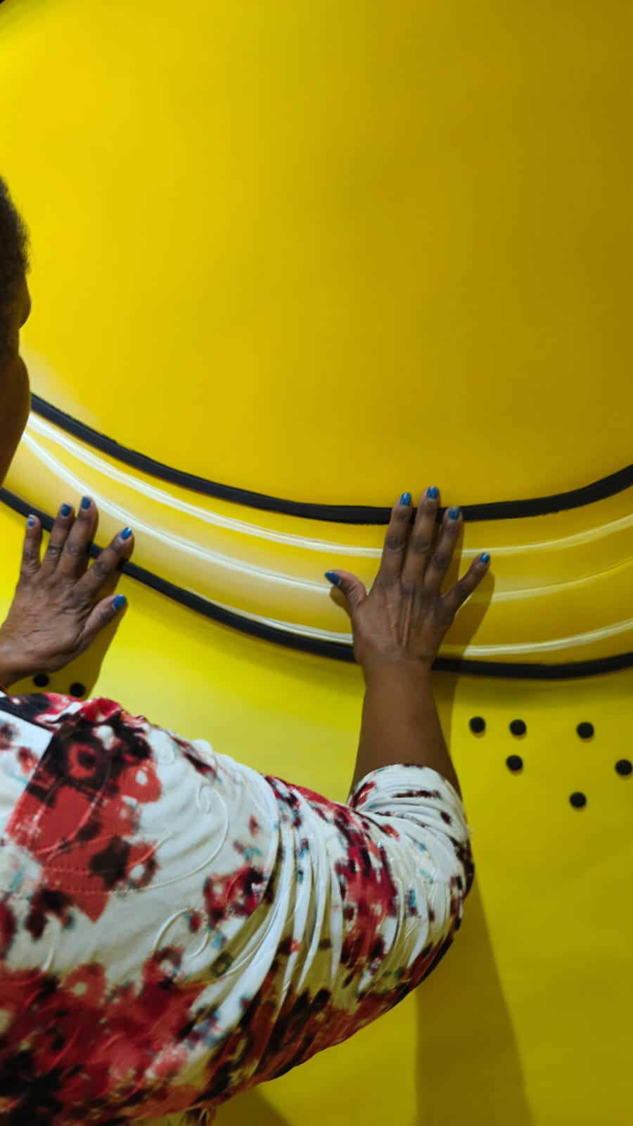 Foto de uma pessoa explorando uma superfície tátil amarela com as mãos. A instalação possui linhas curvas em relevo nas cores preta e branca e pontos em braille na parte inferior. A participante veste blusa estampada em tons vermelhos e mantém as mãos abertas sobre a obra.