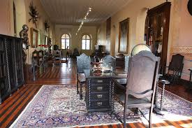 Uma fotografia de plano médio mostra o interior de uma sala histórica em um museu. O ambiente possui um teto alto e plano, paredes em tons de creme com detalhes ornamentais na base e um piso de madeira polida com tábuas largas e escuras. No centro do primeiro plano, há uma escrivaninha de madeira escura com gavetas entalhadas, acompanhada por cadeiras de espaldar alto com detalhes em madeira trabalhada. Sob o conjunto de móveis, estende-se um tapete oriental grande com padrões intrincados em tons de azul, bege e vermelho. Ao longo das paredes, diversos itens históricos estão expostos: À esquerda: Um armário escuro e alto, seguido por uma armadura medieval completa montada em um suporte. À direita: Uma cristaleira de madeira alta com portas de vidro e quadros emoldurados pendurados na parede Ao fundo: Duas janelas em arco permitem a entrada de luz natural, e um lustre trilho moderno no teto ilumina o corredor central da sala. A atmosfera é solene e preservada, evocando um estilo colonial ou clássico europeu.