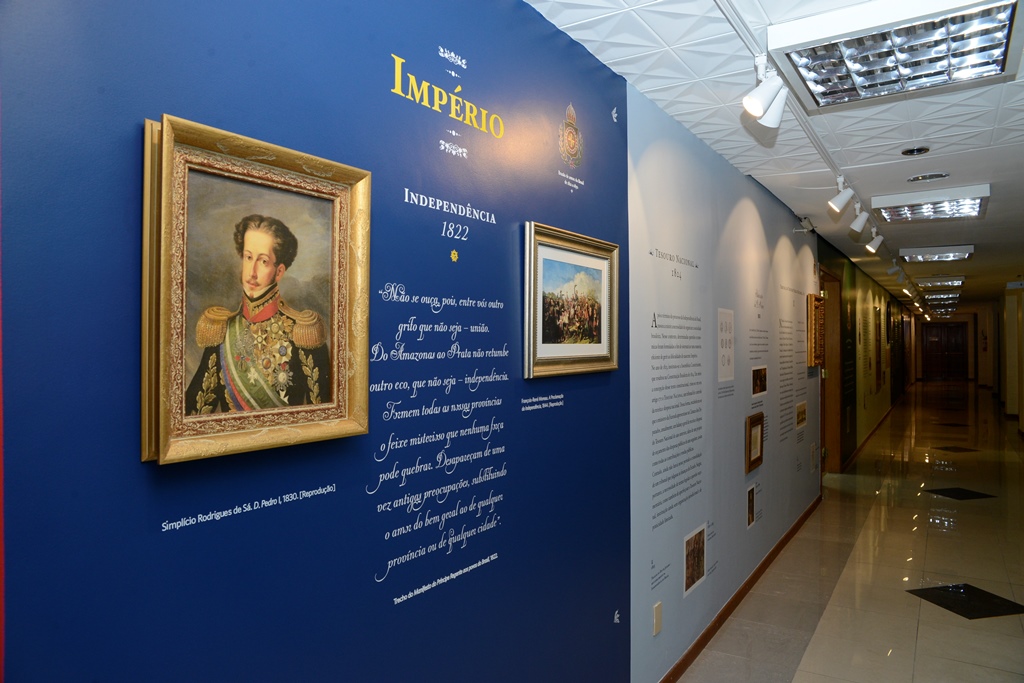 Uma parede com textos e imagens da exposição em fundo azul escuro e depois azul claro. Em destaque vemos uma réplica de retrato do Don Pedro II