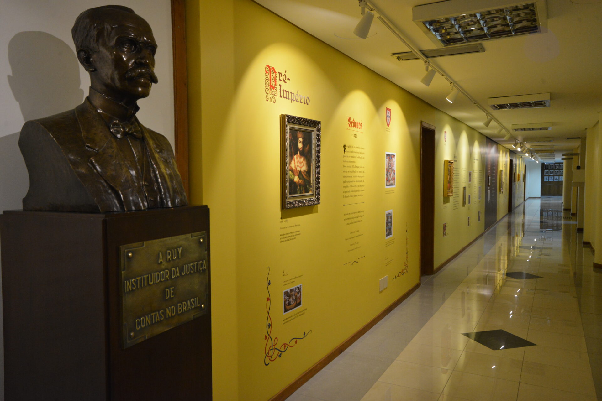 A foto mostra á esquerda um busto de Rui Barbosa e em seguida uma parede com textos e imagens da exposição em fundo amarelo.