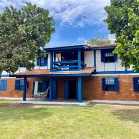 Casa bucólica de dois andares com paredes brancas e detalhes em azul escuro, apresentando seções de tijolo aparente no térreo. Possui janelas com venezianas azuis de madeira maciça, uma área de varanda coberta com colunas de apoio azuis, e uma sacada central no andar superior. O telhado é coberto com telhas de barro vermelho, enquanto árvores frondosas emolduram ambos os lados da propriedade. Um gramado com algumas áreas de grama esparsa se estende na frente da casa sob um céu azul.