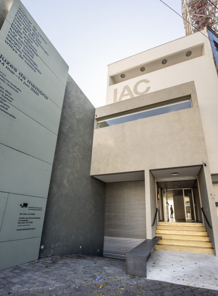 Fotografia em ângulo baixo da fachada externa do Instituto de Arte Contemporânea (IAC). À esquerda, um grande painel cinza-esverdeado exibe uma lista de nomes de artistas e informações sobre a exposição "Luzes da memória", que ocorreu de março a junho de 2020. No centro, a entrada do edifício apresenta uma escadaria curta de madeira que leva a uma porta de vidro aberta, onde se vê a silhueta de uma pessoa ao fundo. Acima da entrada, há uma estrutura de concreto bege com uma janela horizontal estreita e, no topo, a sigla "IAC" em letras grandes e claras. O chão em frente à entrada é pavimentado com pedras escuras e pequenas folhas secas. À direita, no alto, uma parte de uma torre metálica de transmissão é visível contra o céu claro.