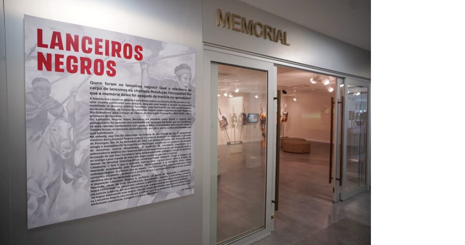Imagem da entrada da exposiçã. à esquerda um painel com o título e texto curatoral. À direita a porta de vidro e a exposição em seu interior.