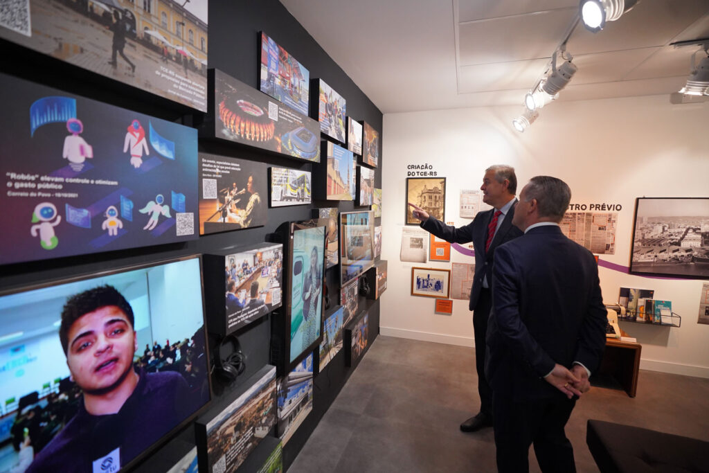 Foto da exposição com uma parede cheia de paineis de notícias sobre o trabalho do tribunal de contas. Duas pessoas olhando e comentando.