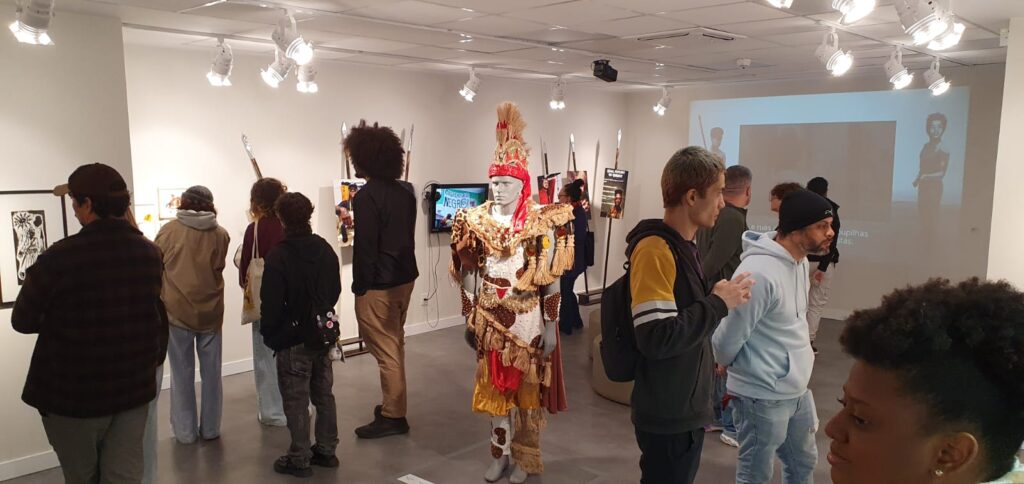Dentro da exposição sobre os Lanceiros Negros, vemos quadros e fotos ao longo das paredes e ao centro um manequim vestindo uma fantasia de carnaval em alegoria sobre Lanceiros Negros. No espaço diversos visitantes observam.