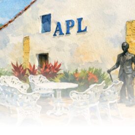 Ilustração artística em aquarela mostrando o pátio externo de um edifício de paredes brancas. Na fachada, destacam-se a sigla "APL" em grandes letras azuis e, mais à direita, uma placa com a inscrição "AUGUSTO DOS ANJOS". Abaixo da placa, há uma estátua escura em tamanho real do poeta vestindo terno e segurando uma bengala. No centro do pátio, encontra-se um conjunto de jardim composto por uma mesa redonda e bancos brancos com detalhes trabalhados. O ambiente é cercado por árvores frondosas à esquerda e folhagens avermelhadas próximas à parede. A borda inferior da imagem possui um efeito suave que desbota para o branco.