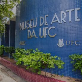 Fachada do Museu de Arte da UFC com seu nome em cor cinza e centralizado em parede azul, com arbustos em sua base.