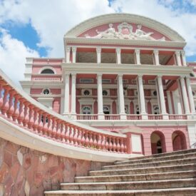Fotografia externa do Teatro Amazonas vista a partir de sua escadaria. Em primeiro plano, destacam-se os degraus de pedra e as curvas dos guarda-corpos com balaústres rosados. Ao fundo, a fachada monumental apresenta colunas brancas, paredes em tons de rosa, arcos, janelas e elementos ornamentais de inspiração clássica. Na parte superior, o frontão decorado se destaca contra o céu azul com nuvens brancas.