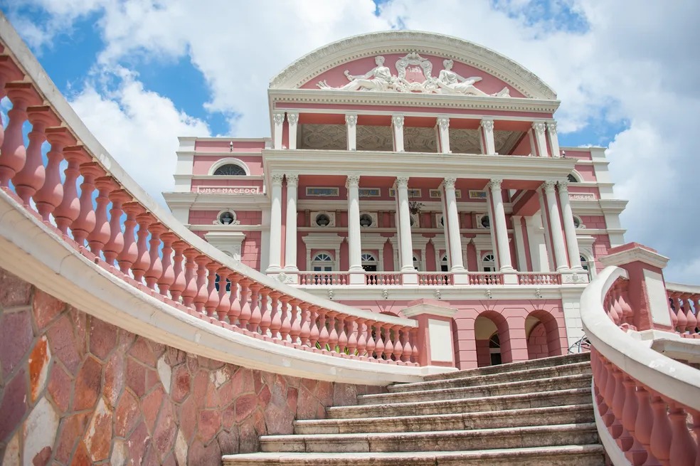 Fotografia externa do Teatro Amazonas vista a partir de sua escadaria. Em primeiro plano, destacam-se os degraus de pedra e as curvas dos guarda-corpos com balaústres rosados. Ao fundo, a fachada monumental apresenta colunas brancas, paredes em tons de rosa, arcos, janelas e elementos ornamentais de inspiração clássica. Na parte superior, o frontão decorado se destaca contra o céu azul com nuvens brancas.