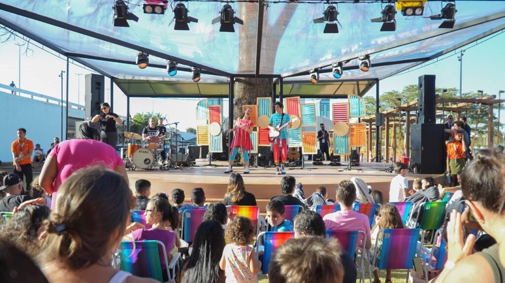Público de crianças e adultos assistindo atração em cadeiras de praia com palco ao fundo.