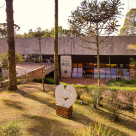 A imagem mostra um jardim com uma escultura branca abstrata. Ao fundo está o Auditório Claudio Santoro, uma construção em formato retangular na cor cinza com janelas de vidro. Ao redor há plantas, árvores.