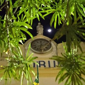 Fotografia noturna mostra a fachada de um prédio histórico com a palavra 'FÓRUM' parcialmente visível. Ao centro, há uma estátua no topo do prédio, acima de um relógio analógico. A lua cheia está visível no céu ao fundo, destacando-se entre as folhas verdes das árvores em primeiro plano. Uma bandeira do Brasil está parcialmente visível na parte inferior da imagem.