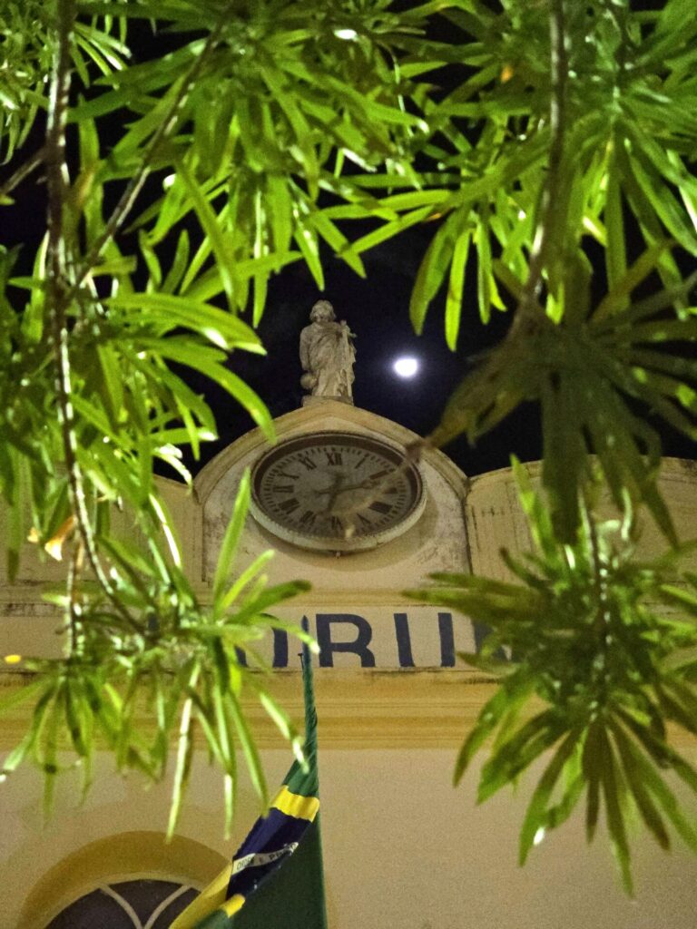 Fotografia noturna mostra a fachada de um prédio histórico com a palavra 'FÓRUM' parcialmente visível. Ao centro, há uma estátua no topo do prédio, acima de um relógio analógico. A lua cheia está visível no céu ao fundo, destacando-se entre as folhas verdes das árvores em primeiro plano. Uma bandeira do Brasil está parcialmente visível na parte inferior da imagem.