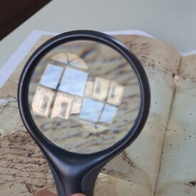 Fotografia mostra uma mão segurando uma lupa sobre um documento antigo com escrita manuscrita. A lupa reflete a imagem de um prédio histórico com janelas arqueadas e detalhes arquitetônicos em pedra. A mesa de fundo é clara, e o documento parece uma carta antiga, parcialmente amarelada e manchada pelo tempo