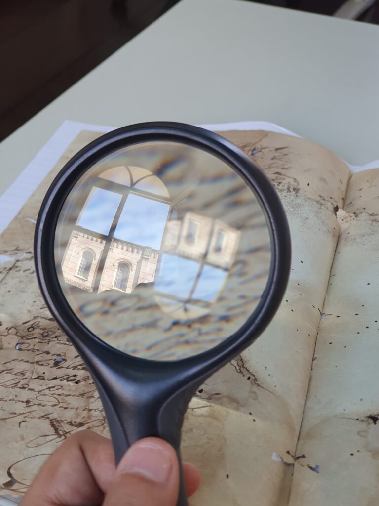 Fotografia mostra uma mão segurando uma lupa sobre um documento antigo com escrita manuscrita. A lupa reflete a imagem de um prédio histórico com janelas arqueadas e detalhes arquitetônicos em pedra. A mesa de fundo é clara, e o documento parece uma carta antiga, parcialmente amarelada e manchada pelo tempo