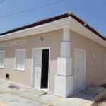 Fachada do Museu Municipal em um prédio térreo de cor bege clara, com detalhes brancos nas portas, janelas e colunas. A entrada principal está aberta, com uma porta dupla branca, acima da qual se lê "MUSEU MUNICIPAL" em letras grandes. O telhado de cerâmica vermelha contrasta com o céu azul claro e sem nuvens. A calçada em frente possui piso tátil para acessibilidade.