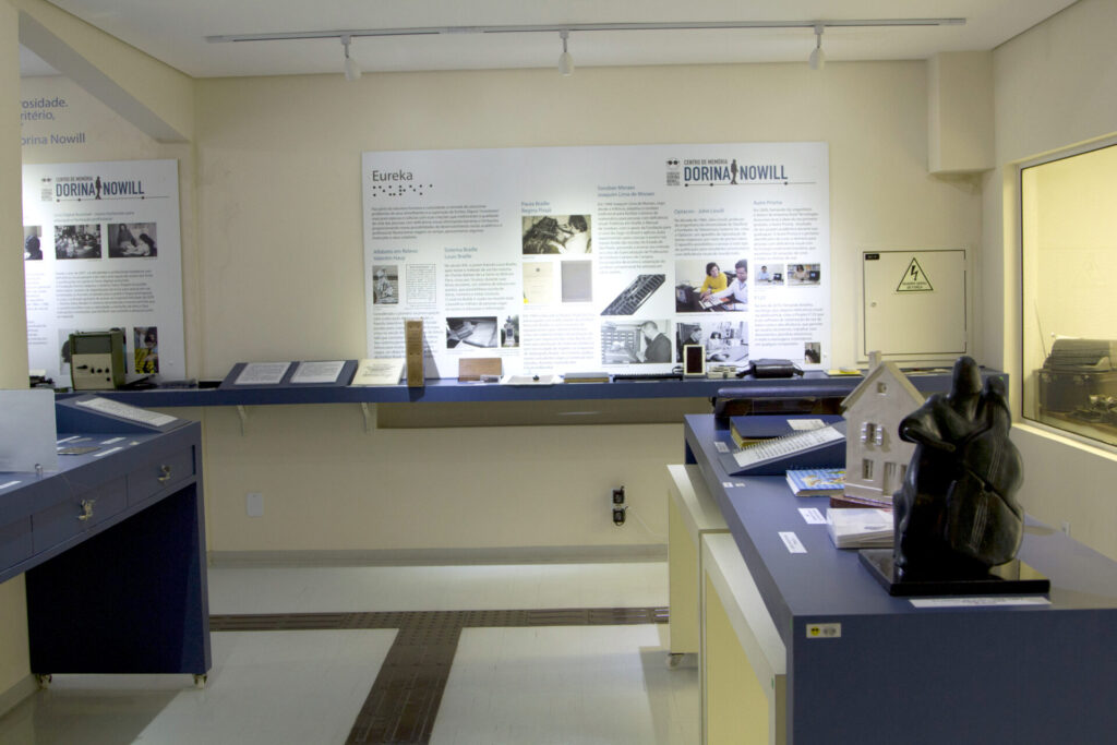 Fotografia do Centro de Memória da Fundação Dorina Nowill para Cegos. O ambiente é iluminado e exibe painéis expositivos na parede com textos, imagens e informações sobre a história da Fundação e do Sistema Braille. Sobre mesas azuis, há objetos históricos como livros em braille, equipamentos de leitura e escrita tátil, uma maquete branca de uma casa e uma escultura escura em forma de figura humana abraçada a um violoncelo. No chão, piso tátil direcional e de alerta auxilia a locomoção de pessoas cegas e com baixa visão pelo espaço.