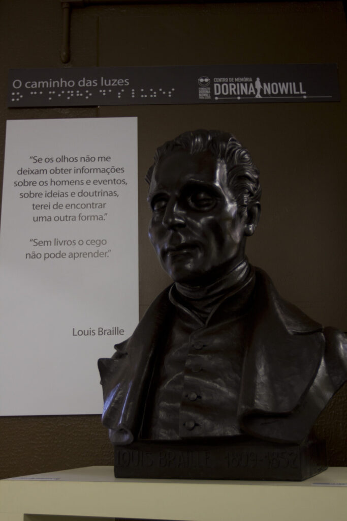 Fotografia de um busto em bronze de Louis Braille, criador do sistema de leitura e escrita para pessoas cegas. A escultura está exposta sobre uma base clara no Centro de Memória da Fundação Dorina Nowill para Cegos. Atrás, na parede marrom, há um painel com a frase “O caminho das luzes” em letras e em braille. Ao lado da escultura, um painel branco exibe a citação de Louis Braille: “Se os olhos não me deixam obter informações sobre os homens e eventos, sobre ideias e doutrinas, terei de encontrar uma outra forma. Sem livros o cego não pode aprender.”