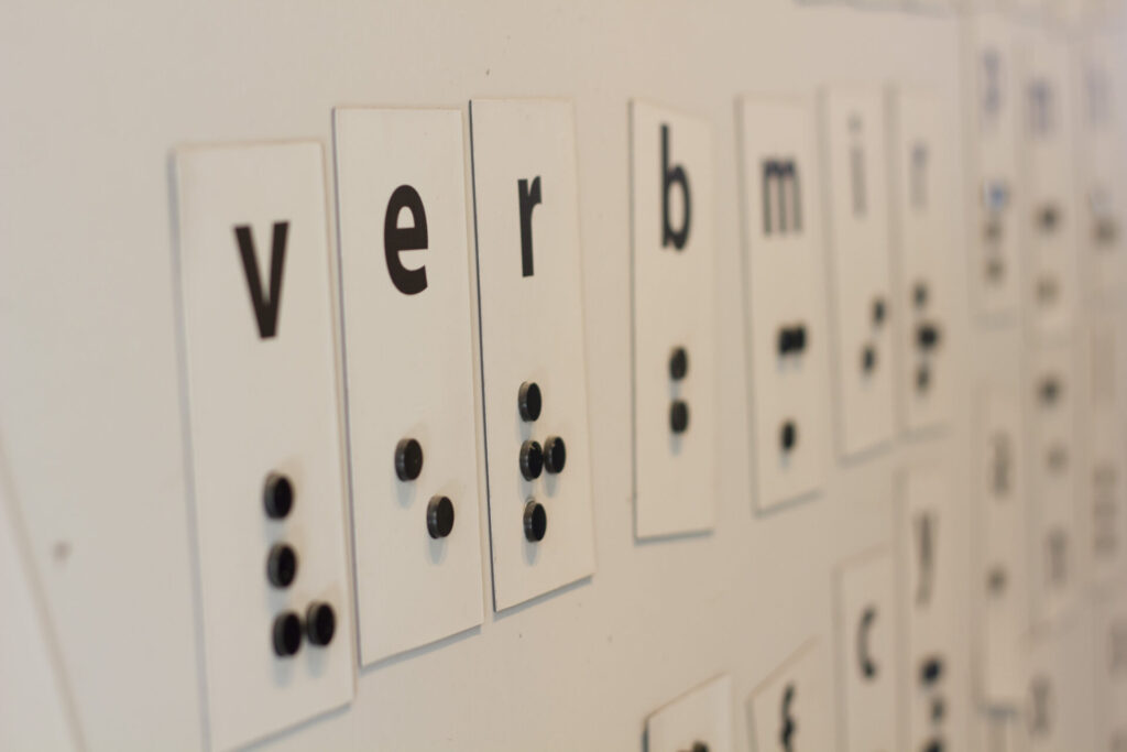 Placas brancas alinhadas em uma parede exibem letras do alfabeto latino acompanhadas de suas respectivas representações em braille, feitas com pequenos pinos pretos em relevo. Na imagem, destacam-se as letras "v", "e", "r" e "b". A cena faz parte de uma exposição educativa sobre o sistema braille, permitindo a visualização simultânea da grafia comum e da tátil.