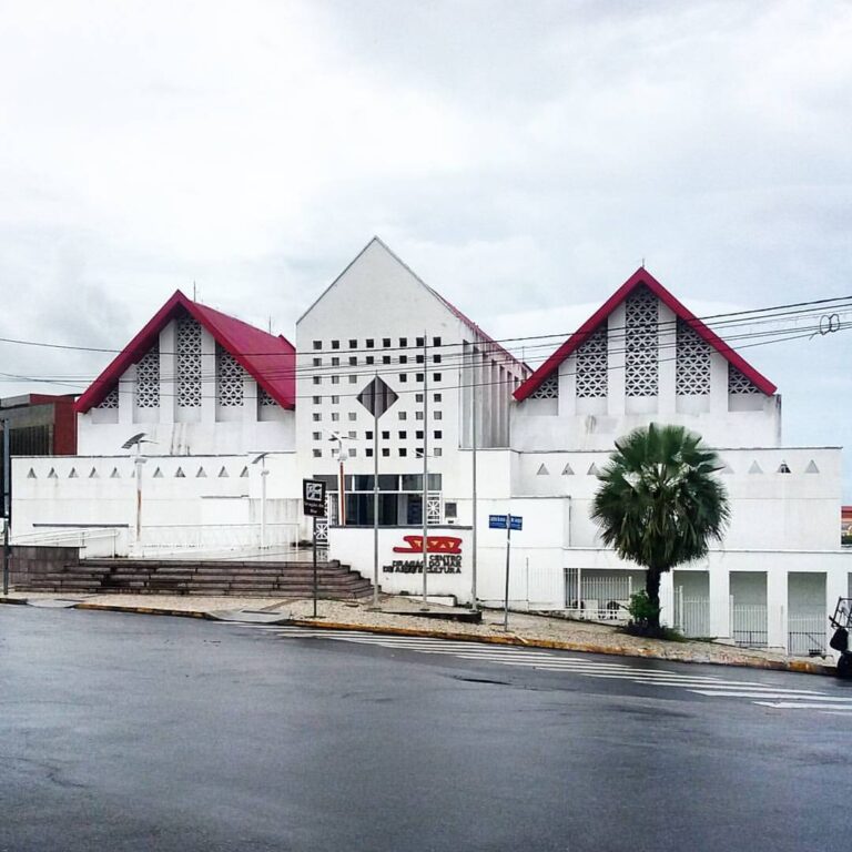 A imagem mostra a fachada de um edifício de arquitetura moderna. Sua estrutura é composta por três telhados em formato triangular. A fachada principal é branca com detalhes em vermelho visíveis nos telhados dos prédios da extremidade esquerda e direita. Há painéis com elementos vazados geométricos nas fachadas. No prédio do centro um detalhe vazado em formato de losango, com espaços quadrados ao redor, criando padrões decorativos. No centro há uma larga escadaria de cinco degraus. A direita, na mureta, a logo do Centro Dragão do Mar de Arte e Cultura, nas cores vermelha e amarela.