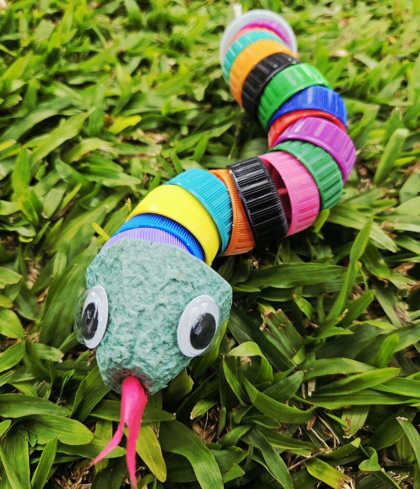 Cobra artesanal confeccionada com tampinhas plásticas coloridas reutilizadas, com olhos móveis e língua bifurcada rosa, fotografada sobre a grama.