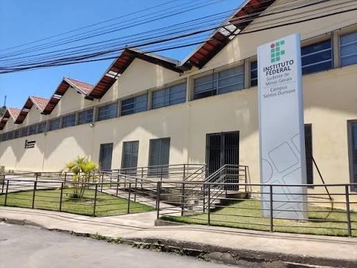 A fachada do Instituto Federal do Sudeste de Minas Gerais - Campus Santos Dumont em um dia ensolarado. A edificação possui uma arquitetura industrial ou acadêmica de linhas retas, com paredes pintadas em tom bege claro e um telhado serrilhado característico (estilo "shed") com telhas cerâmicas visíveis. Na frente do prédio, há um totem vertical branco com o logotipo verde e vermelho da instituição e a inscrição "INSTITUTO FEDERAL Sudeste de Minas Gerais Campus Santos Dumont". O acesso à entrada principal conta com rampas e escadas protegidas por corrimãos metálicos escuros, cercados por pequenas áreas de gramado e um arbusto decorativo, com o céu azul limpo ao fundo e fiação elétrica cruzando o topo da cena.