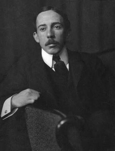 A imagem apresenta um retrato fotográfico em preto e branco do inventor Alberto Santos-Dumont da cintura para cima, posicionado em um plano médio. Ele aparece sentado e voltado ligeiramente para a direita, com o olhar direcionado fixamente para a câmera em uma expressão séria e concentrada. Santos-Dumont veste um traje formal escuro, composto por paletó e colete, combinados com uma camisa social de colarinho alto e rígido na cor branca, além de uma gravata escura volumosa. O punho de sua camisa branca se projeta sob a manga esquerda do paletó, exibindo uma abotoadura. Ele tem cabelos escuros penteados para trás com risca lateral, olhos expressivos e um bigode farto bem aparado sobre os lábios. A iluminação destaca suavemente o seu rosto e as partes brancas da vestimenta contra o fundo escuro e plano.