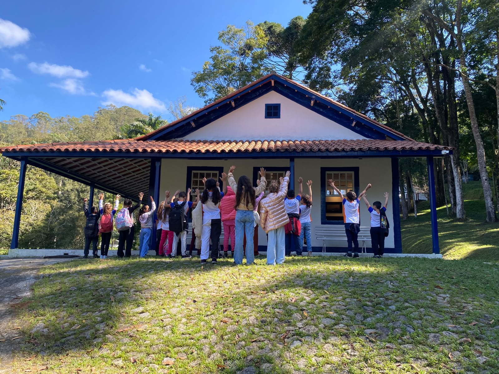 A imagem mostra um grupo de crianças de costas em frente à casa branca de janelas azuis, todos com os braços levantados para o alto. Eles vestem roupas coloridas e casacos, e alguns usam mochilas. O grupo está na varanda da casa, que fica em cima de um gramado verde com um caminho de pedras na frente. Ao redor, há árvores altas e um céu azul com sol.