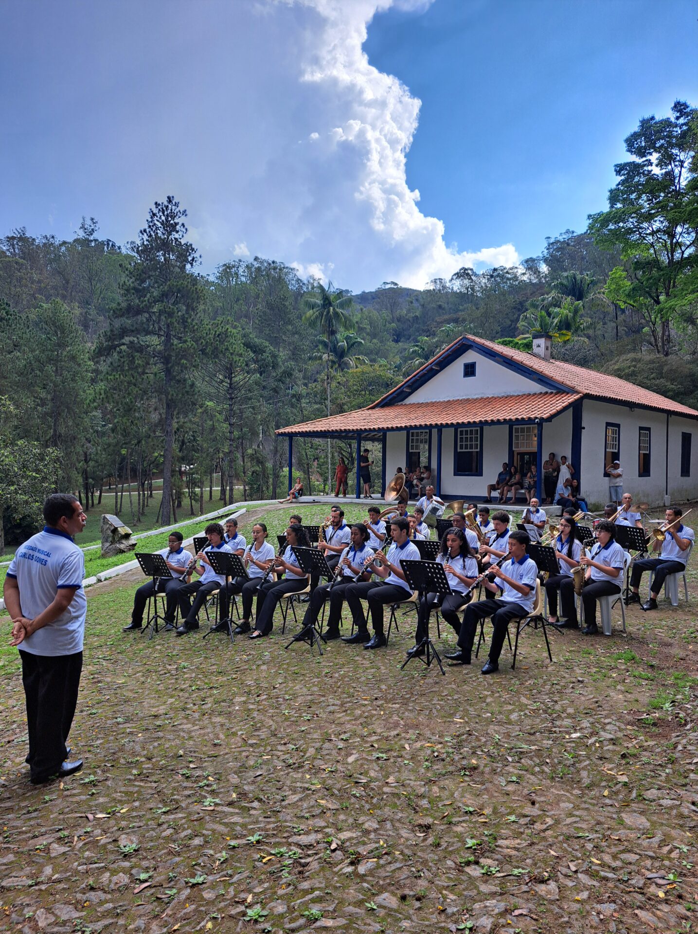 A imagem mostra uma banda musical de jovens se apresentando sentados ao ar livre em frente à mesma casa branca com detalhes azuis. Os músicos, que usam camisas brancas e calças pretas, estão dispostos em fileiras com seus instrumentos de sopro e partituras em pedestais. Em primeiro plano, à esquerda e de costas, um maestro de camisa branca observa o grupo. O cenário ao redor é composto por um chão de pedras irregulares, um gramado verde, árvores altas nas laterais e montanhas ao fundo, sob um céu azul com uma grande nuvem branca.