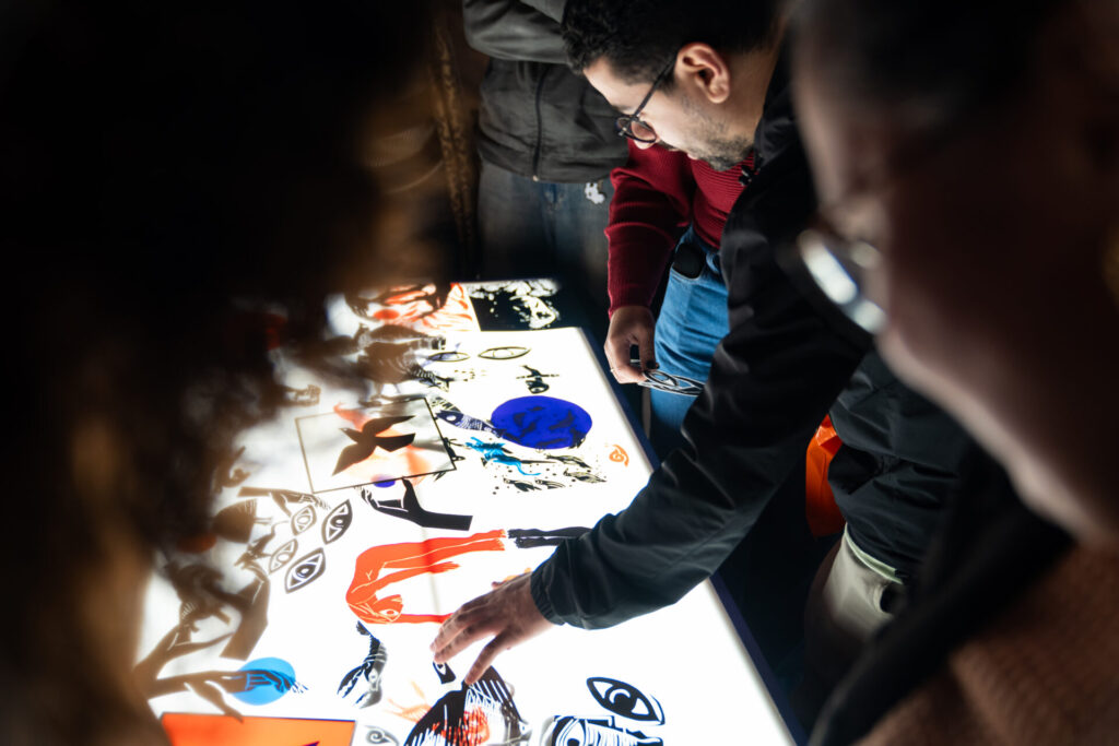 Visitantes interagem com uma mesa de luz, sobre a qual estão dispostas imagens e formas gráficas relacionadas às obras da exposição. A fotografia registra uma atividade participativa e de aproximação do público com o universo visual do artista.