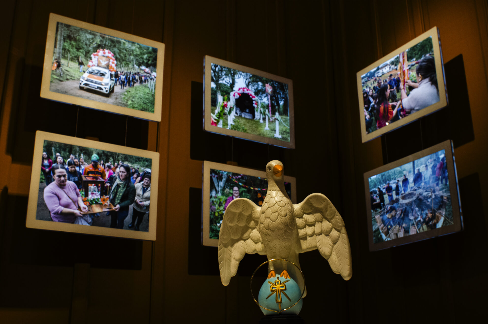Fotografia com iluminação em tom quente e suave, em ambiente escuro, destacando uma escultura branca em primeiro plano que representa um pombo de asas abertas sobre uma esfera azul-clara adornada com uma auréola e uma cruz douradas. Ao fundo, dispostas em formato de arco nas paredes escuras de uma galeria, encontram-se cinco telas emolduradas em branco que exibem fotografias coloridas da exposição, mostrando pessoas reunidas em celebrações ao ar livre, procissões com carros decorados e rituais comunitários.