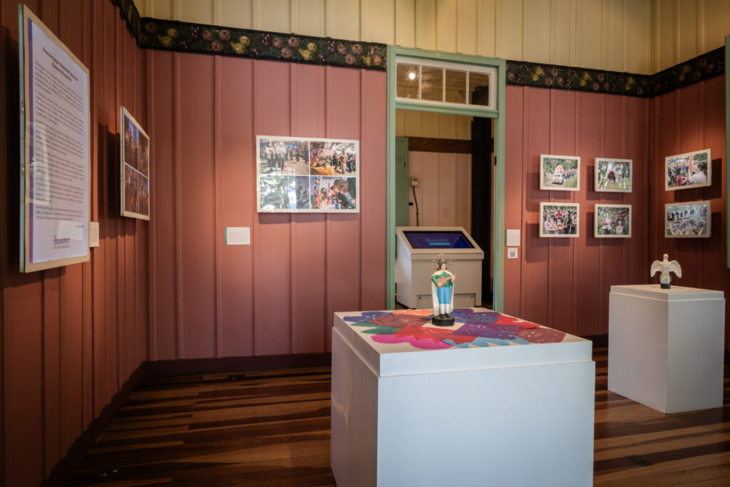 Fotografia de ambiente interno da Casa Domingos Nascimento Sobrinho, sede do IPHAN-PR em Curitiba, com piso de madeira escura e paredes de madeira pintadas em rosa antigo, com um friso floral decorativo superior. A sala abriga a exposição "Povos do Campo: Patrimônio Cultural da Floresta de Araucárias". Em primeiro plano, à esquerda, vê-se um grande painel de texto vertical branco com título e corpo de texto em português. Ao centro, uma escultura de santo de pé, vestindo túnica branca e manto verde, sobre uma base de papel picado colorido em formato de flor, repousa em um pedestal branco. À direita, um segundo pedestal branco exibe uma pequena escultura de pombo branco sobre esfera azul e cruz dourada. Nas paredes, diversos conjuntos de fotografias emolduradas e painéis de texto menores documentam as comunidades rurais e indígenas. Ao fundo, uma porta emoldurada em verde-claro leva a outra sala, onde um quiosque interativo com tela azul é visível. A iluminação é artificial, focada nos objetos e painéis. A fotografia é de autoria de Douglas Fróis.