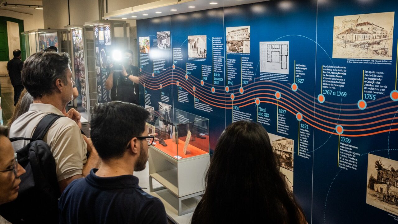 Fotografia em plano médio com iluminação interna e tom geral azulado, mostrando visitantes de costas observando um grande painel azul na Sede Histórica do MAE-UFPR em Paranaguá. O painel exibe uma linha do tempo detalhada com textos, fotografias históricas, documentos e ilustrações conectados por linhas sinuosas laranja e branca, cobrindo o período desde o século XVIII (1755) até os anos recentes. Abaixo do painel, há vitrines expositoras de vidro com peças museológicas. Em primeiro plano, o perfil de três pessoas de cabelo escuro e óculos acompanha atentamente os dados históricos, enquanto no reflexo e ao fundo aparecem outros visitantes e a estrutura da galeria. A fotografia é de autoria de Douglas Fróis.