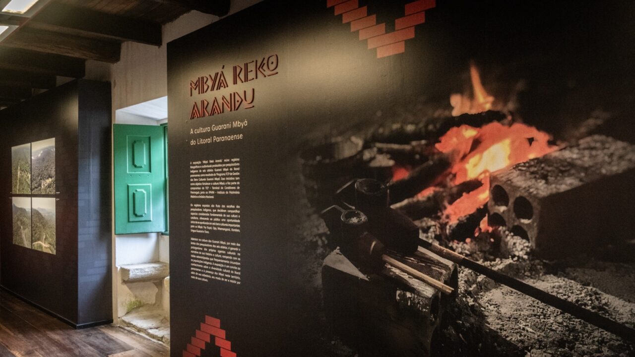 Fotografia em plano médio com iluminação interna quente, focada em um painel expositivo preto da exposição "Mbyá Rekó Arandu" no MAE-UFPR. No centro superior do painel, lê-se em letras grandes e brancas: "MBYÁ REKÓ ARANDU" e, logo abaixo, "A cultura Guarani Mbyá do Litoral Paranaense". Abaixo, um longo texto explicativo detalha a mostra. À direita, uma projeção brilhante exibe imagens de um fogo crepitante e um braseiro sobre pedras, com um bloco de concreto e ferramentas de metal próximas, remetendo a rituais. À esquerda, um nicho em uma parede branca abriga duas fotografias emolduradas de paisagens montanhosas e verdes, com uma porta de madeira pintada de verde-azulado ao lado. O chão é de madeira e o teto exibe vigas de madeira escura. A fotografia é de autoria de Douglas Fróis.