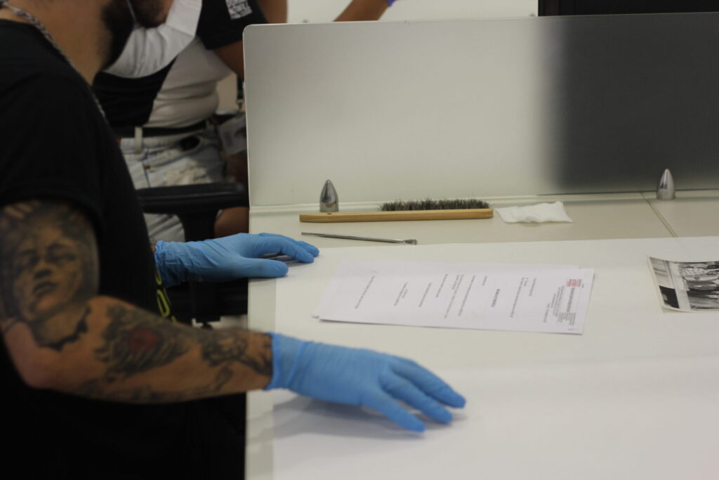 A imagem mostra uma pessoa com luvas azuis manipulando documentos em uma mesa branca. Há uma divisória metálica, um pincel, uma ferramenta metálica e uma fotografia preta e branca. A pessoa tem tatuagens no braço esquerdo.