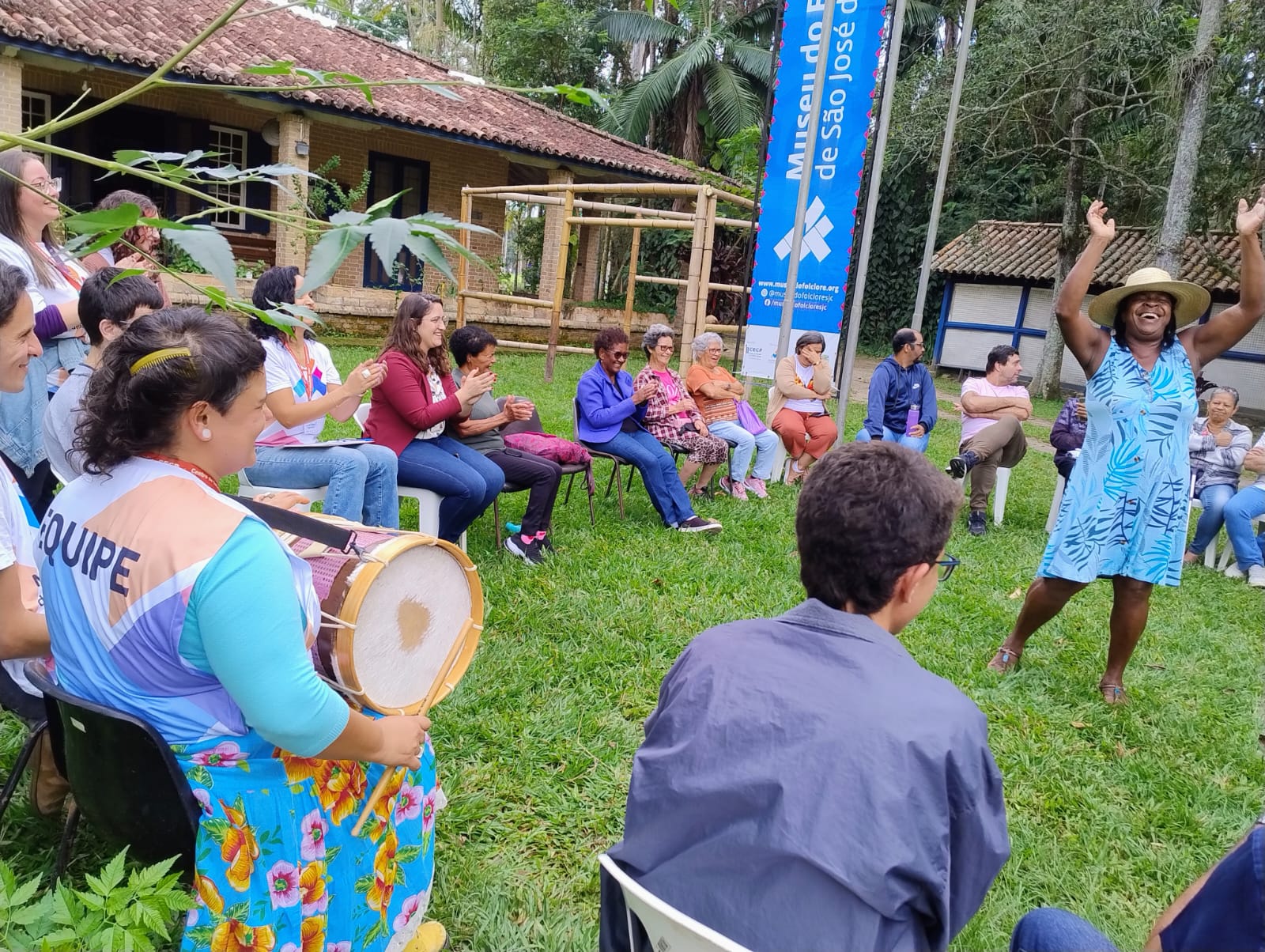 Pessoas de várias etnias sentadas em roda na frente do Museu do Folclore. Dentro da roda uma mulher negra sorrindo e de braços para cima.