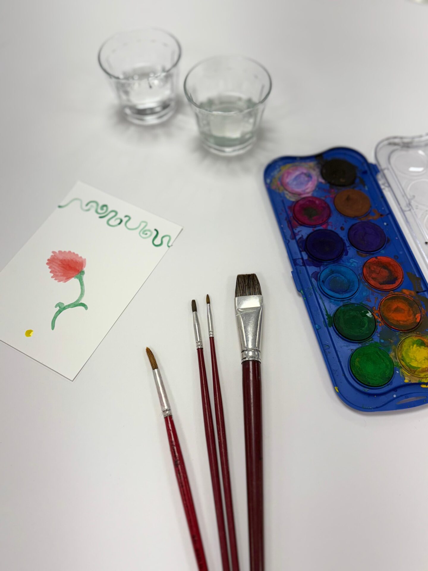 Um estojo de aquarela, quatro pincéis, dois copos de água para limpar os pincéis e um trabalho em aquarela